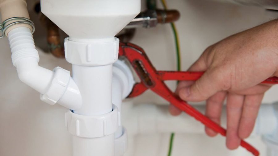 Person using a red pipe wrench to tighten PVC plumbing under a sink, close-up view