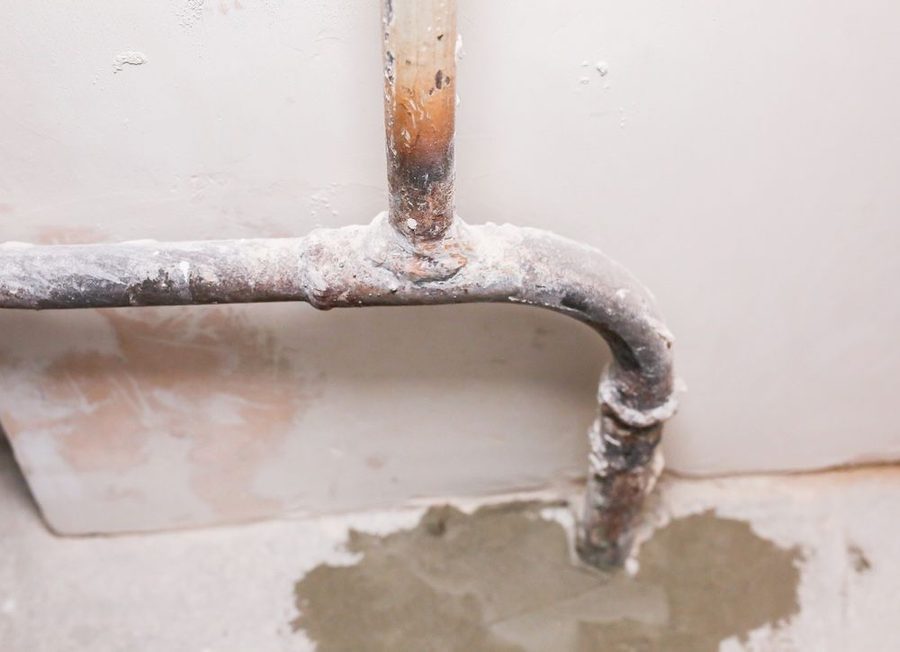 Old, rusted metal pipe with joint, against a wall with plaster damage and white corrosion buildup